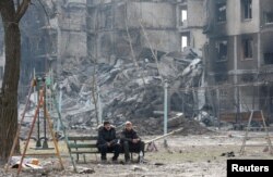 Penduduk setempat duduk di bangku dekat sebuah gedung apartemen yang hancur akibat konflik Ukraina-Rusia di kota pelabuhan selatan yang terkepung Mariupol, Ukraina, 25 Maret 2022. (Foto: REUTERS/Alexander Ermochenko)