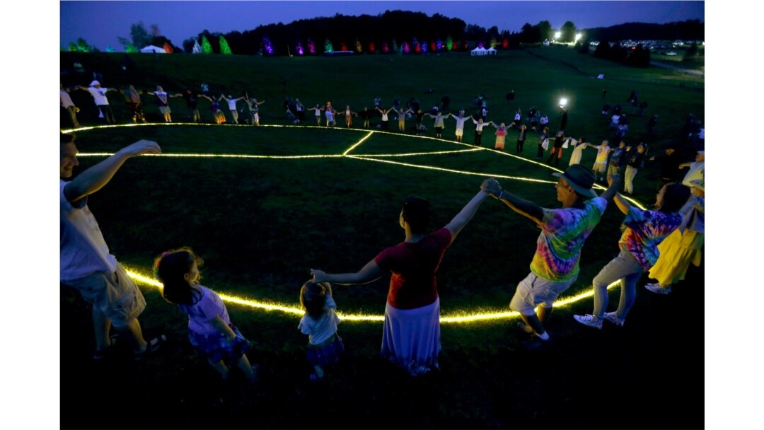 People hold hands in a circle around a large, illuminated peace sign on the original site of the 1969 Woodstock Music and Arts Fair in Bethel, N.Y., Aug. 15, 2019.