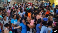 n this Aug. 30, 2019, photo, migrants, many who were returned to Mexico under the Trump administration’s “Remain in Mexico,” program wait in line to get a meal in an encampment near the Gateway International Bridge in Matamoros.