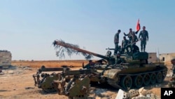 In this Aug. 13, 2019 photo, released by the Syrian official news agency SANA, Syrian army soldiers flash the victory sign as they stand on a tank, in northwestern Syria. 