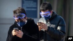 Travelers wear face masks and goggles as they use their smartphones outside the Beijing Railway Station in Beijing, Saturday, Feb. 15, 2020. (AP Photo/Mark Schiefelbein)