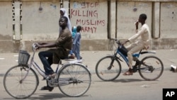 Dua remaja naik sepeda di kota Kano, Nigeria dengan tembok bertuliskan "Stop Membunuh Muslim, Tentara". Tentara Nigeria dituduh membantai Muslim Syiah di Kaduna dan menguburnya secara massal. 