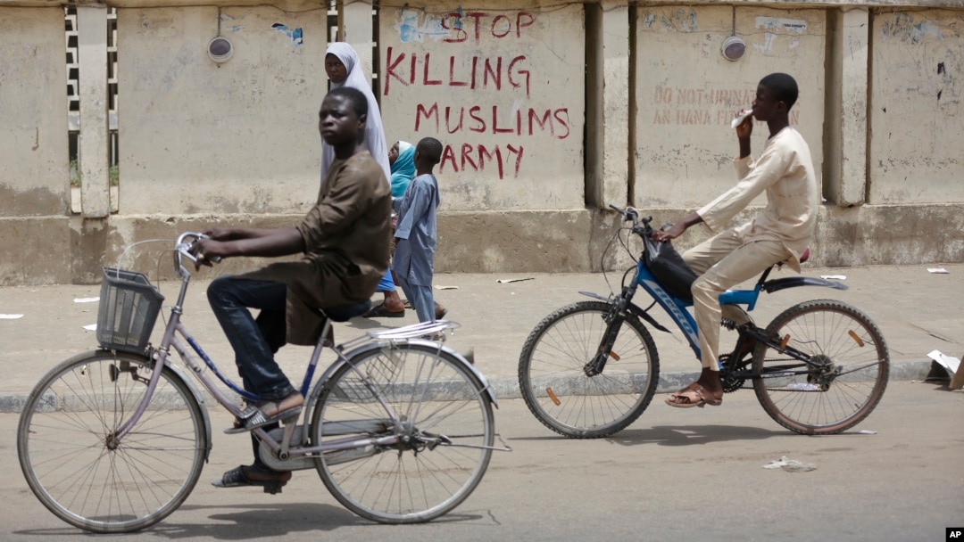 Dua remaja naik sepeda di kota Kano, Nigeria dengan tembok bertuliskan "Stop Membunuh Muslim, Tentara". Tentara Nigeria dituduh membantai Muslim Syiah di Kaduna dan menguburnya secara massal. 