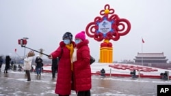 Women wearing face masks printed with a slogan for the Beijing Winter Olympics Games take a selfie with a decoration for the Winter Olympics Games in Tiananmen Square in Beijing on Jan. 20, 2022.