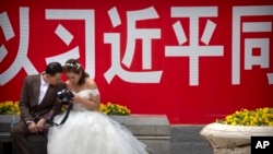 资料照：一对穿着婚礼服装的夫妇2018年5月19日在北京观看照相机背面的照片，身后是关于中国国家主席习近平的宣传口号。