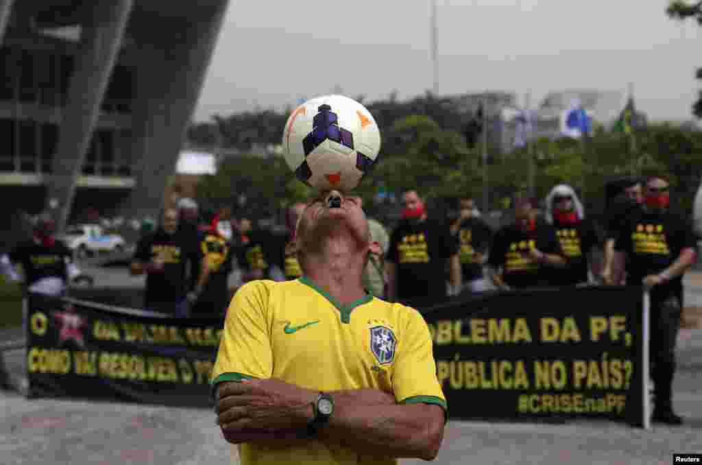 Um fã de futebol domina a bola com a cabeça, enquanto agentes da polícia federal protestam por melhores salários e condições de trabalho, atrás dele. Brasil 2014