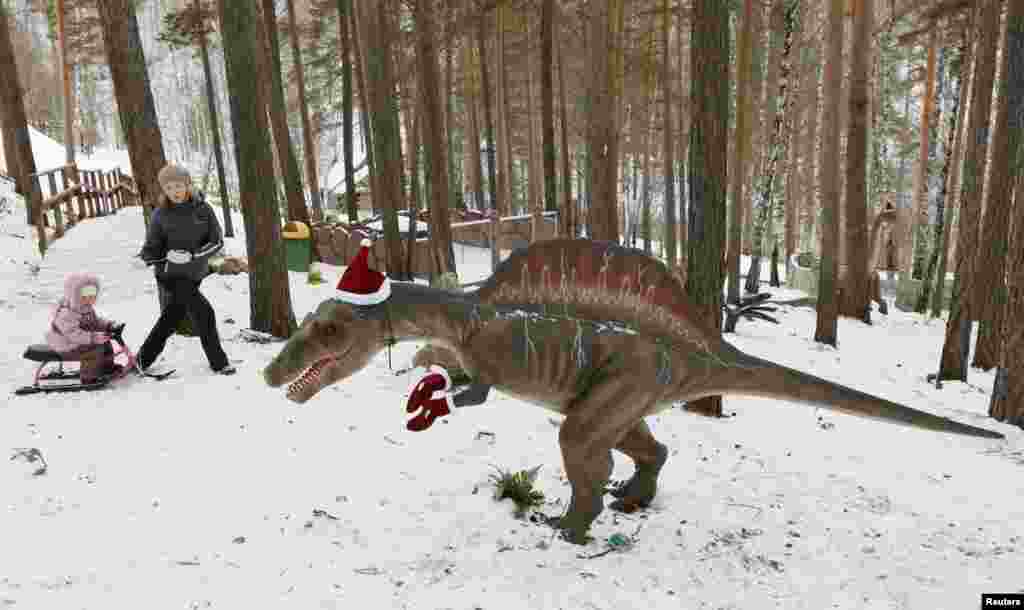 People walk past a model of a dinosaur, with Santa Claus hat and gloves on it, at the Dinosaurs Park located in the Taiga area at the Royev Ruchey zoo in the suburbs of Russia&#39;s Siberian city of Krasnoyarsk.