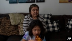 Marina Maalouf, a longtime resident of Hillside Villa, sits on a sofa as her granddaughter eats pizza for lunch in their apartment in Los Angeles, Oct. 1, 2024.