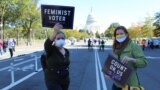 Duas mulheres fotografadas com o capitólio ao fundo exibem cartazes a dizer "Eleitora Feminista" e "Conta conosco para votar". Marcha das Mulheres, 17 de outubro 2020