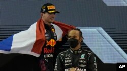 Lewis Hamilton no pódio, enquanto Max Verstappen passa por trás depois da vitória no Grande Prémio de ABu Dhabi. 12 Dezembro 2021