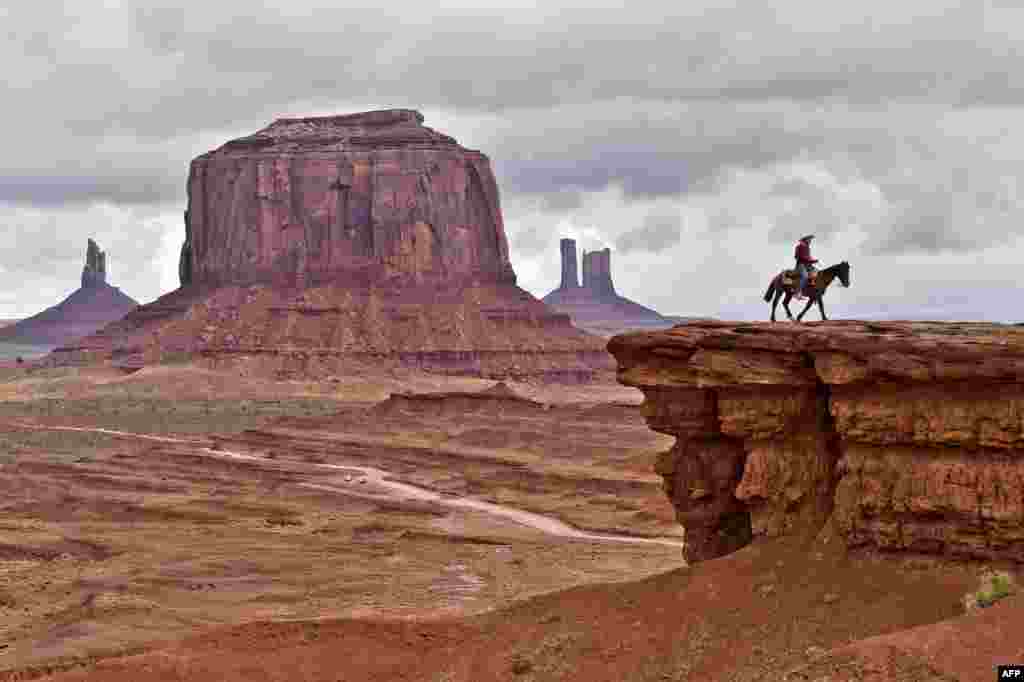 បុរសកុលសម្ព័ន្ធ Navajo ថតរូបពេលជិះសេះ នៅខាងមុខកូនភ្នំ Merrick នៅក្នុងឧទ្យាន Monument Valley Navajo Tribal Park រដ្ឋ Utah សហរដ្ឋអាមេរិក។