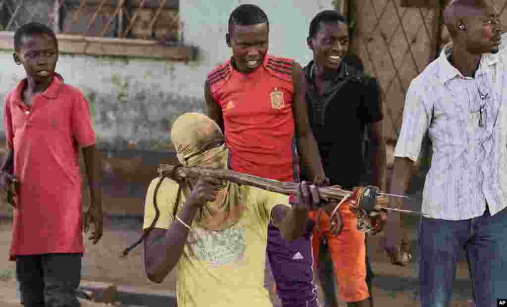 Một người biểu tình che mặt,&nbsp;ở quận Buyenzi của thủ đô Bujumbura, Burundi, chĩa chiếc gậy, với những con dao cột vào đầu gậy, hướng về phía cảnh sát. Những người biểu tình cho biết họ sẽ tiếp tục phản đối cho đến khi Tổng thống Pierre Nkurunziza rời chức vào cuối nhiệm kỳ thứ hai của ông.