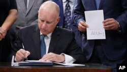 Gov. Jerry Brown signs a copy of the environmental measure SB100 as the bill's author, state Sen. Kevin de Leon (R) holds a copy of the bill, Sept. 10, 2018, in Sacramento, California. 