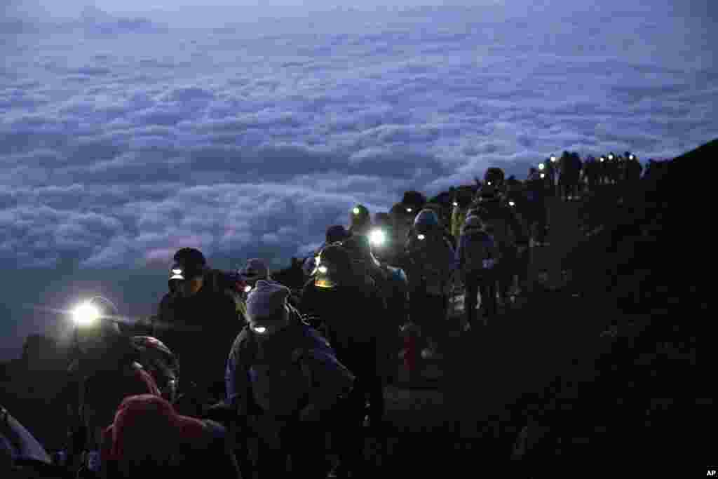 A group of hikers climb to the top of Mount Fuji, Japan, just before sunrise as clouds hang below the summit.