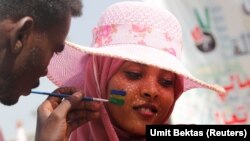 A Sudanese protester waits for a man to finish a painting of the Sudan's old national flag on her face