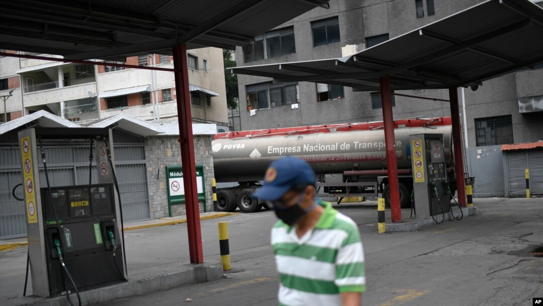 Un hombre con un tapabocas camina frente a una estación de servicio de una compañía petrolera estatal vacía mientras un camión cisterna descarga gasolina en Caracas.