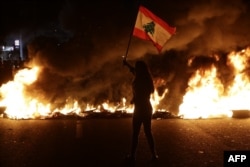 A Lebanese anti-government protester waves a Lebanese flag in front of burning tires that block the main highway linking the city of Tripoli to Beirut at the coastal city of Byblos, north of the capital, Nov. 13, 2019.