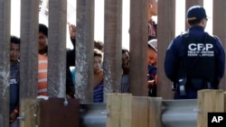 ARSIP – Sekumpulan kaum migran dari Amerika Tengah memandang melalui sela-sela dinding perbatasan saat petugas U.S. Customs and Border Protection, kanan, bersiaga di sisi yang berlawanan, Minggu, 25 November 2018 (foto: AP Photo/Gregory Bull)