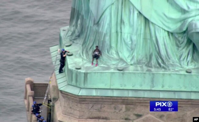 In this image made from video by WPIX, Therese Okoumou, center, leans against the robes of the Statue of Liberty on Liberty Island, as a police officer stands on a ledge nearby, talking her into descending, in New York, July 4, 2018.
