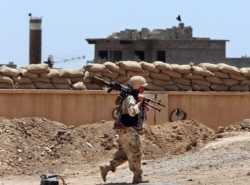FILE - A Kurdish peshmerga fighter carries his weapon as he walks to his base where two Islamic State flags are seen on a building, right, and a water tower, left, south of Kirkuk, northern Iraq, June 25, 2014.