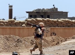 FILE - A Kurdish peshmerga fighter carries his weapon as he walks to his base where two Islamic State flags are seen on a building, right, and a water tower, left, south of Kirkuk, northern Iraq, June 25, 2014.