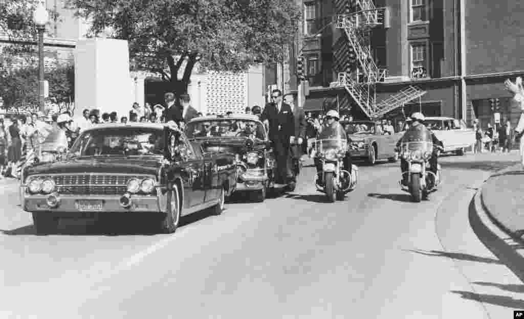 Seen through the limousine's windshield as it proceeds along Elm Street past the Texas School Book Depository, President John F. Kennedy appears to raise his hand toward his head within seconds of being fatally shot in Dallas, Nov 22, 1963. 