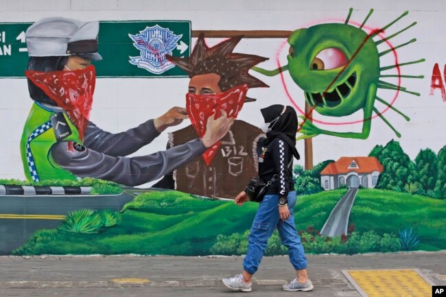 Seorang perempuan berjalan melewati mural bertema virus corona yang menyerukan agar masyarakat selalu menggunakan masker untuk menekan penyebaran COVID-19, di Medan, Sumatera Utara, Indonesia, Senin 1 November 2021. (Foto: AP)