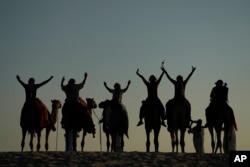 People pose for a photo atop camels in Mesaieed, Qatar, Nov. 26, 2022. Throngs of World Cup fans in Qatar looking for something to do between games are leaving Doha for a classic Gulf tourist experience: riding a camel in the desert. But the sudden rise in tourists is putting pressure on the animals, who have almost no time to rest between each ride. (AP Photo/Ashley Landis)