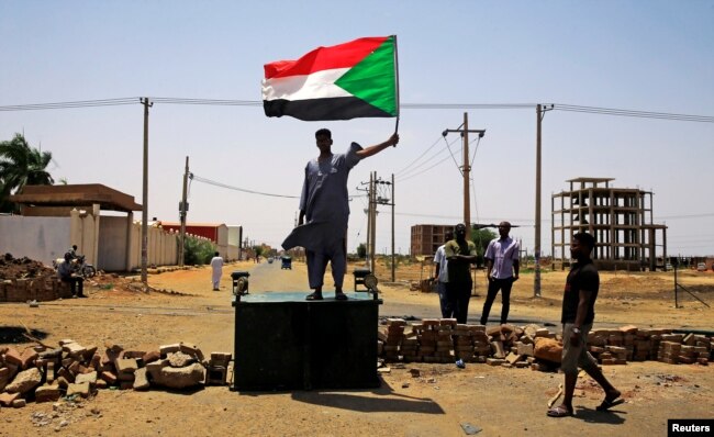 FILE - A Sudanese protester holds a national flag as he stands on a barricade along a street, demanding that the country's Transitional Military Council hand over power to civilians, in Khartoum, Sudan, June 5, 2019.