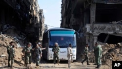 FILE - In this photo released on April 30, 2018 by the Syrian official news agency SANA, Syrian government forces gather in front of a bus carrying al-Qaida-linked fighters during an evacuation from the Palestinian refugee camp 0f Yarmuk, near Damascus. 