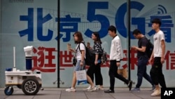 People walk toward a police robot mounted with surveillance cameras patrol past a 5G network advertisement at a shopping district in Beijing, May 15, 2019. 