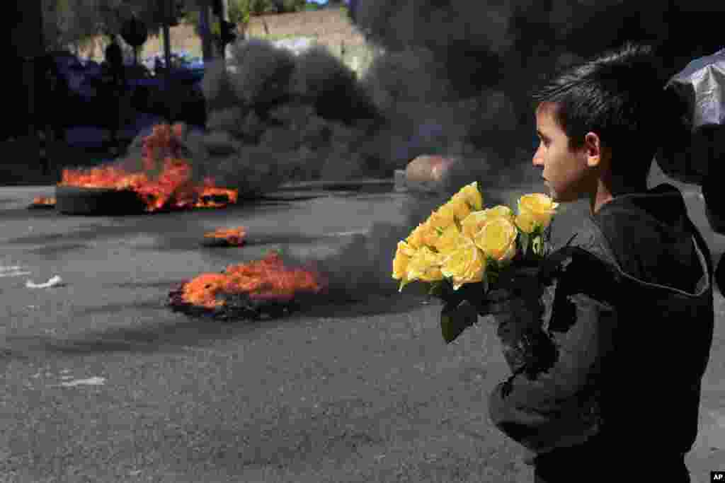A Syrian boy who sells flowers on the street watches protesters burn tires to block a main road during a protest against the increase in prices of consumer goods and the crash of the local currency, in Beirut, Lebanon.