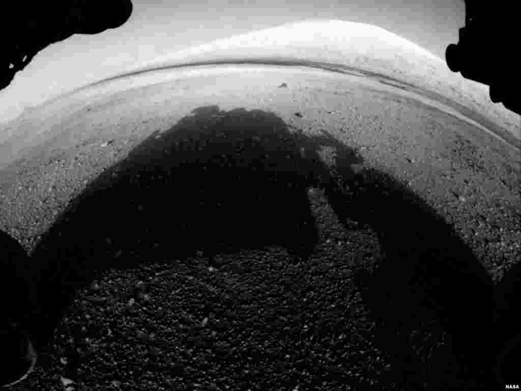 This image shows what lies ahead for the rover -- its main science target, informally called Mount Sharp. The rover&#39;s shadow is seen in the foreground, and the dark bands beyond are dunes. In the distance is the highest peak of Mount Sharp.