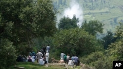 Smoke rises behind from a mortar shell fired by Pakistani troops as Indian villagers sit during the funeral of Indian civilian Sarpanch Karamat Hussain who was killed in Pakistani shelling at Balakot sector in Poonch, Jammu and Kashmir, India, Aug.16, 201