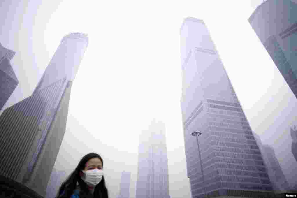 Một người phụ nữ đeo khẩu trang đi bên dưới những tòa nhà chọc trời giữa khói mùa tại khu tài chính Phố Đông, Thượng Hải, Trung Quốc. Cảnh báo ô nhiễm màu đỏ đầu tiên từ trước tới giờ của Trung Quốc đối có hiệu lực từ đầu tuần này.