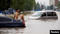 一名男子坐在暴雨后郑州街头漂浮的汽车上。（2021年7月22日）