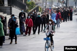 A woman wearing a face mask rides a shared bicycle past people lining up to enter a supermarket in Wuhan, Hubei province, the epicentre of China's coronavirus disease (COVID-19) outbreak, April 1, 2020.