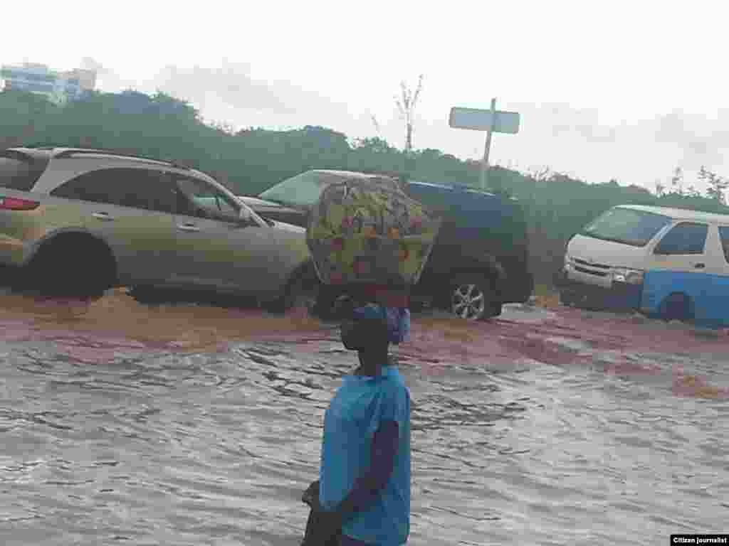 Mulher caminhando com trouxa à cabeça por rua inundada em Luanda