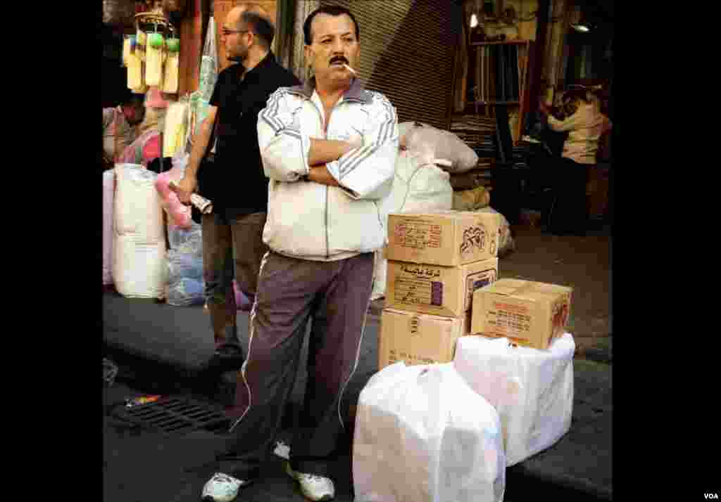 A man waits on the side of the street in a Damascus market with boxes full of goods. (J. Weeks/VOA)