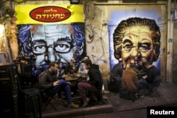 People dine near portraits of Albert Einstein (L) and former Israeli Prime Minister Golda Meir (R), spray-painted on metal shutters of closed storefronts in Mahane Yehuda, one of Jerusalem's most popular outdoor markets, Feb. 24, 2016.