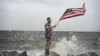 El youtuber Mark Peyton sostiene una bandera estadounidense mientras posa para su hermano Matt Peyton en la costa antes de la llegada del huracán Helene a Alligator Point, Florida, el 26 de septiembre de 2024.