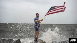 El youtuber Mark Peyton sostiene una bandera estadounidense mientras posa para su hermano Matt Peyton en la costa antes de la llegada del huracán Helene a Alligator Point, Florida, el 26 de septiembre de 2024.