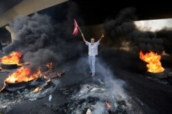 An anti-government protester makes victory sign, as he holds a Lebanese national flag and walks fire of tires that sits to block a road during a protest against government's plans to impose new taxes in Beirut, Lebanon, Oct. 18, 2019.