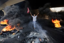 An anti-government protester makes victory sign, as he holds a Lebanese national flag and walks fire of tires that sits to block a road during a protest against government's plans to impose new taxes in Beirut, Lebanon, Oct. 18, 2019.