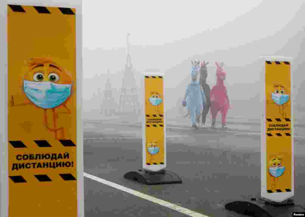 People wearing costumes walk near a skating rink and past signs urging the use of protective face masks and to keep a social distance amid the COVID-19 outbreak, as heavy fog covers Lenin Square in Stavropol, Russia.