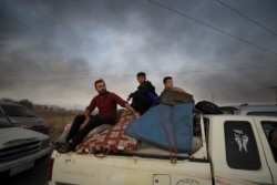 A man and two boys sit on belongings at the back of a truck as they flee Ras al-Ayn, Syria, Oct. 9, 2019, with smoke billowing in the background during a Turkish offensive.