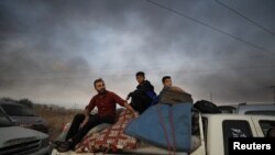 A man and two boys sit on belongings at the back of a truck as they flee Ras al-Ayn, Syria, Oct. 9, 2019, with smoke billowing in the background during a Turkish offensive.