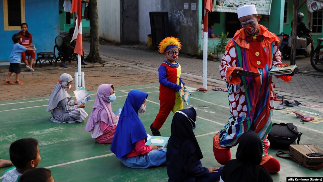 Yahya Edward Hendrawan, guru berusia 38 tahun yang berpakaian badut untuk memimpin kelas, ditemani putranya Mirza yang berusia 5 tahun, berpakaian seperti bayi badut, membagikan buku kepada anak-anak saat mengajar di sebuah desa selama bulan puasa suci Ra