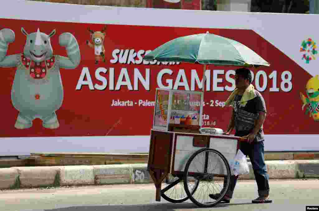 Seorang pedagang makanan mendorong gerobaknya di sebuah jalan di Jakarta, melewati spanduk promosi Asian Games yang akan dilaksanakan di Jakarta dan Palembang, 15 Juli 2018. (Foto: Reuters)