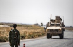 FILE - A fighter from the Syrian Democratic Forces stands guard as a convoy of U.S. military vehicles drives on a road after U.S. forces pulled out of their base in the northern Syrian town of Tal Tamr, Oct. 20, 2019.